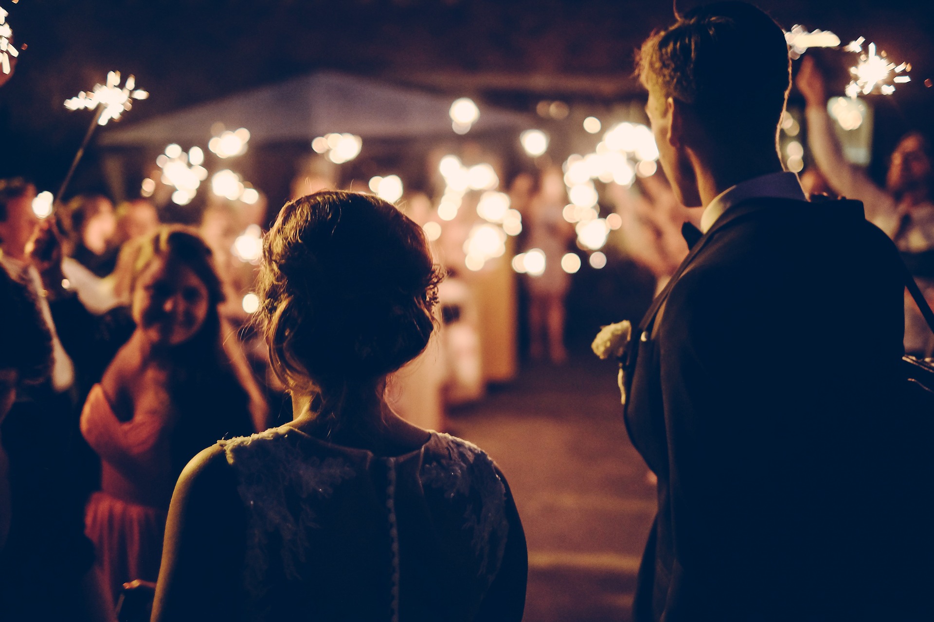 A bride and groom staring at a woman in a pink dress at a wedding. The woman in the pink dress is welcoming with a bright smile. It's evening, and the lighting is dim, provided by string lights.